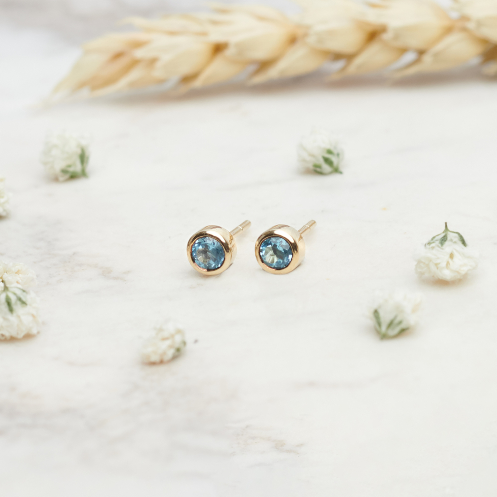 Tiny Swiss Blue Topaz Earrings in Solid Gold on a background with flowers