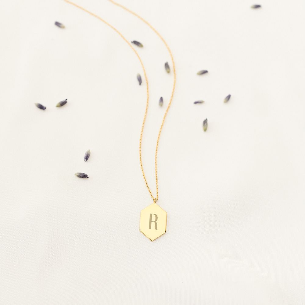 A Hexagon Pendant Necklace with an Engraved Monogram in yellow Gold laid down on a white sheet with leaves surrounding it.